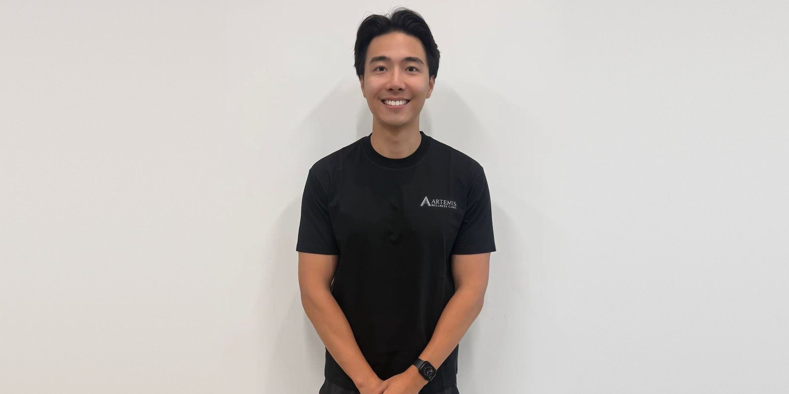 Artemis Wellness Clinic ਦੇ ਰਜਿਸਟਰਡ ਫਿਜ਼ੀਓਥੈਰੇਪਿਸਟ Colin, ਕਲੀਨਿਕ ਦੀ ਕਾਲੀ ਟੀ-ਸ਼ਰਟ ਪਹਿਨੇ, ਹਲਕੇ ਰੰਗ ਦੀ ਕੰਧ ਅੱਗੇ ਮੁਸਕਰਾਉਂਦੇ ਹੋਏ