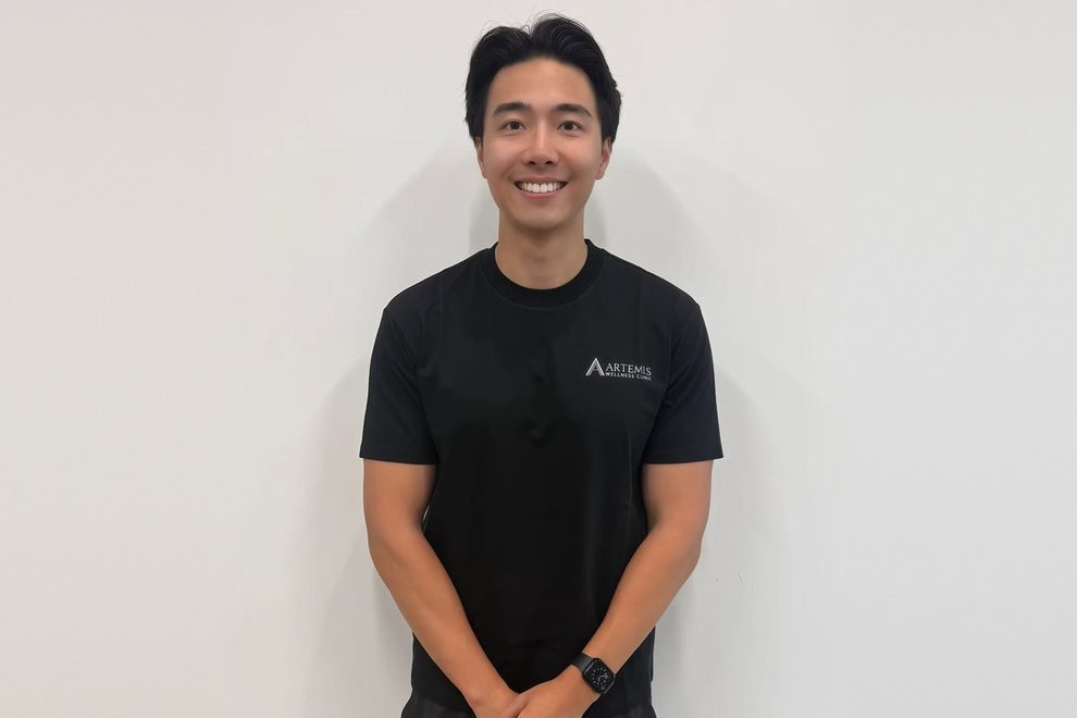 Artemis Wellness Clinic ਦੇ ਰਜਿਸਟਰਡ ਫਿਜ਼ੀਓਥੈਰੇਪਿਸਟ Colin, ਕਲੀਨਿਕ ਦੀ ਕਾਲੀ ਟੀ-ਸ਼ਰਟ ਪਹਿਨੇ, ਹਲਕੇ ਰੰਗ ਦੀ ਕੰਧ ਅੱਗੇ ਮੁਸਕਰਾਉਂਦੇ ਹੋਏ